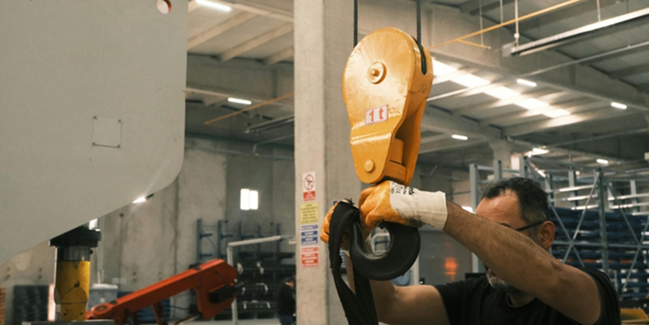 A worker wearing safety gloves operates an orange industrial hoist in a spacious, well-lit warehouse.