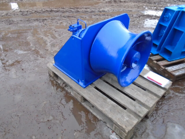 A large, shiny blue industrial capstan winch sat on a wooden pallet in a muddy outdoor area.