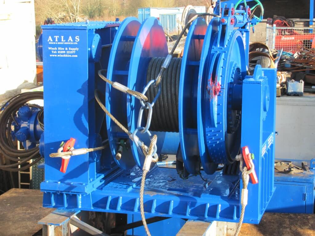 A large blue industrial winch is shown outdoors, with metal cables coiled around it.