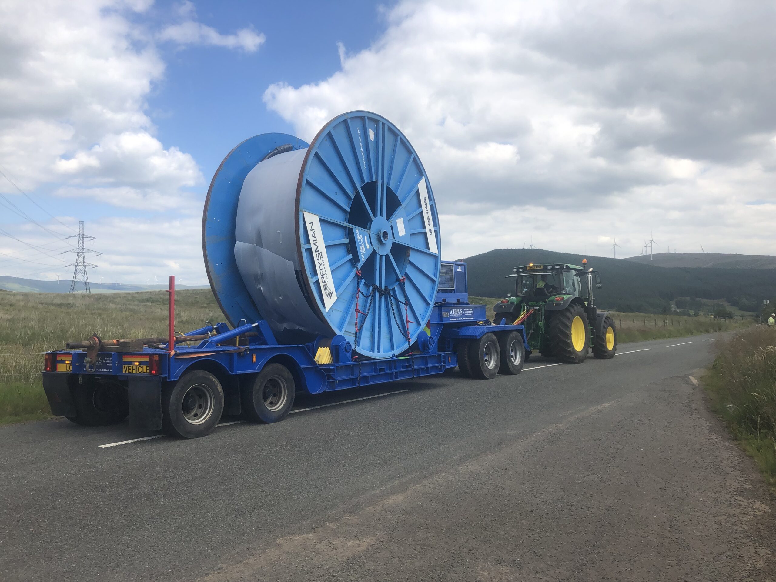 A large blue cable drum on a trailer is being pulled by a green tractor on an open road.