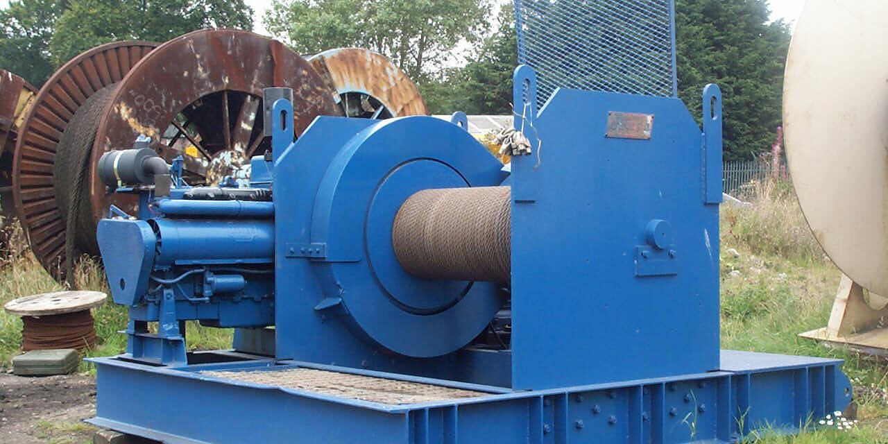 A large, industrial blue winch sits outdoors on a concrete surface. It features a thick metal cable coiled around its drum.