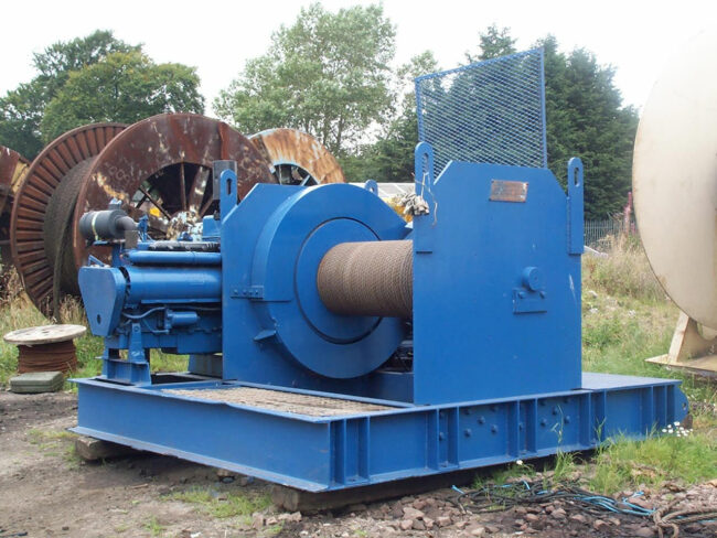 A large, industrial blue winch sits outdoors on a concrete surface. It features a thick metal cable coiled around its drum.