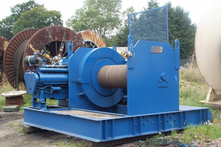 A large, industrial blue winch sits outdoors on a concrete surface. It features a thick metal cable coiled around its drum.