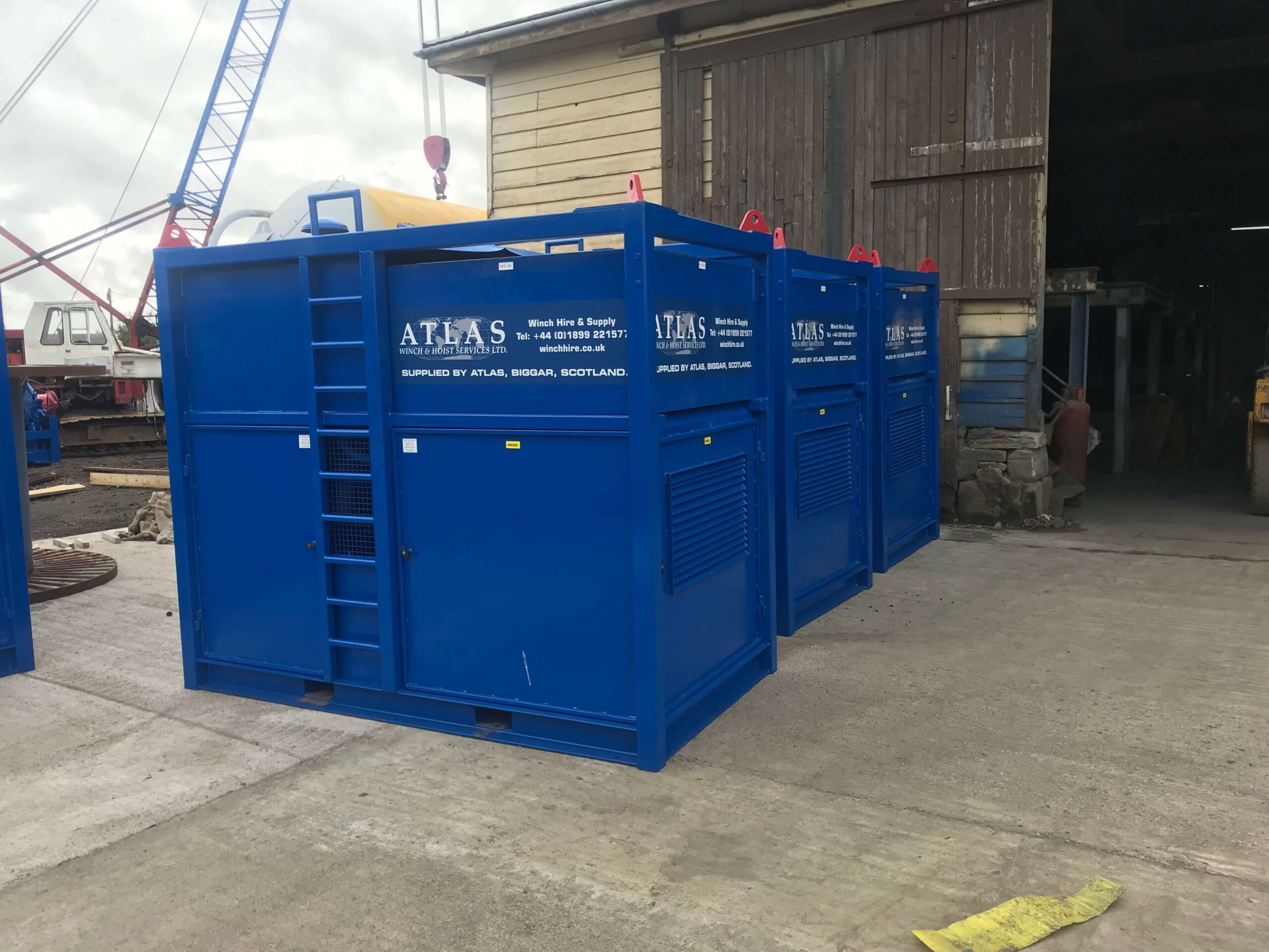 Blue industrial equipment containers labeled "ATLAS" are lined up outside a large wooden warehouse.