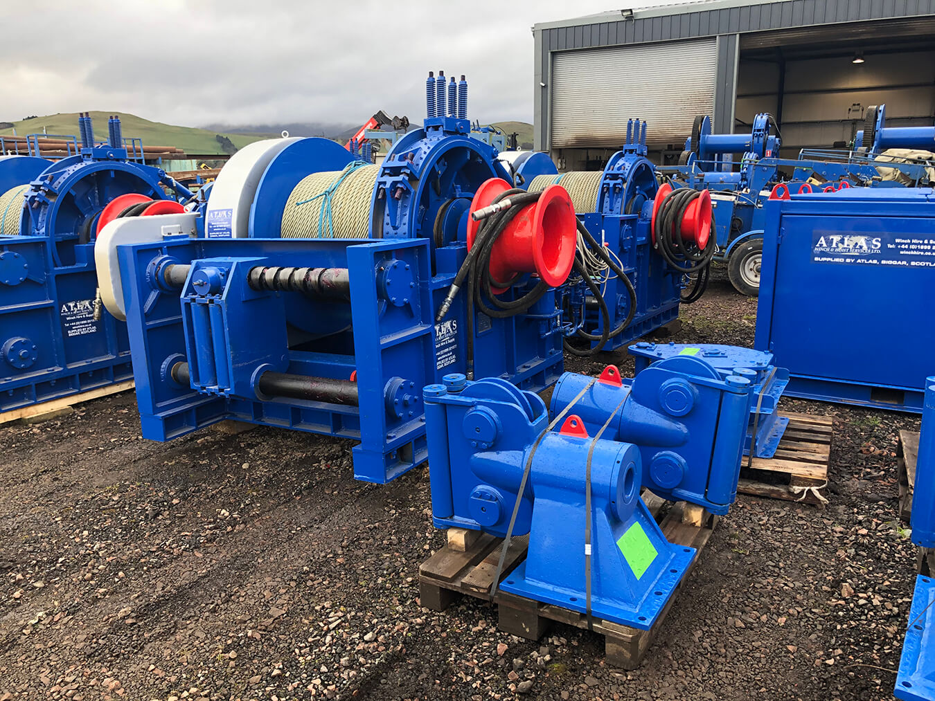 Blue industrial winches on a gravel lot outside a warehouse with coiled ropes and red hooks.