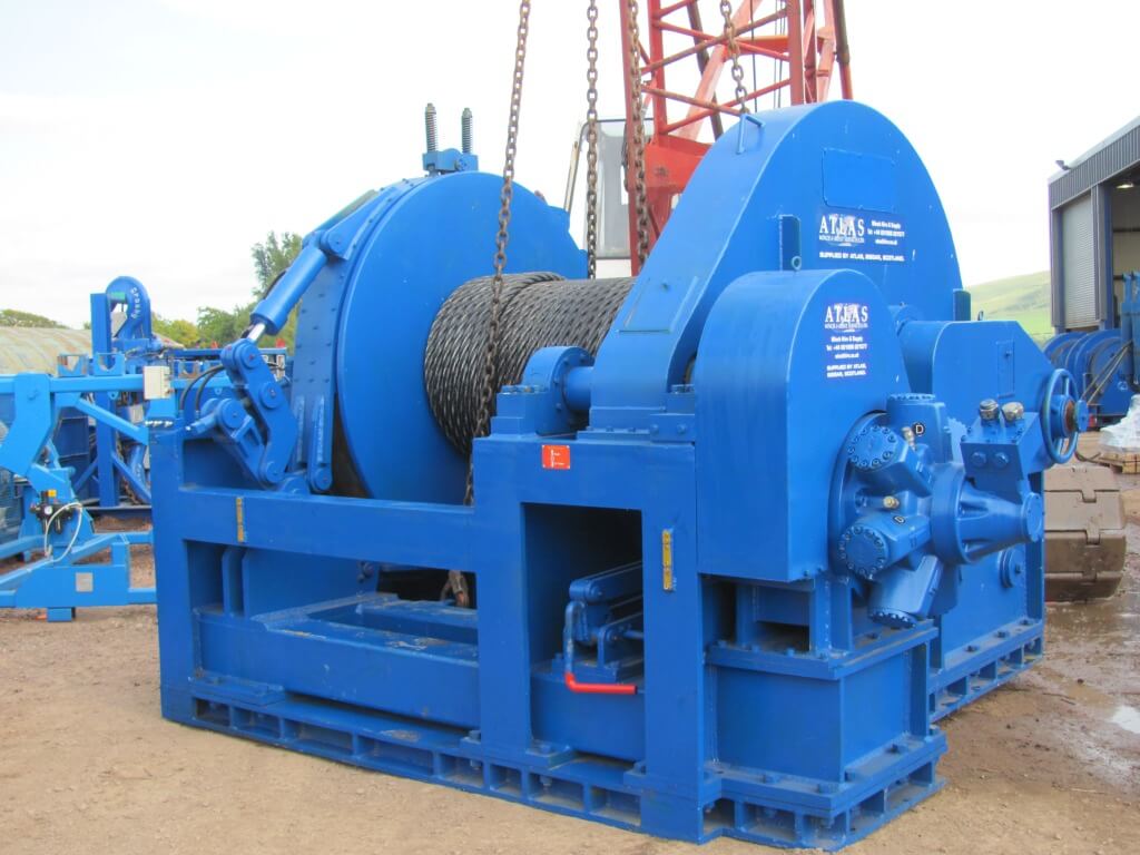Industrial blue winch with coiled rope, situated outdoors with a rural backdrop.