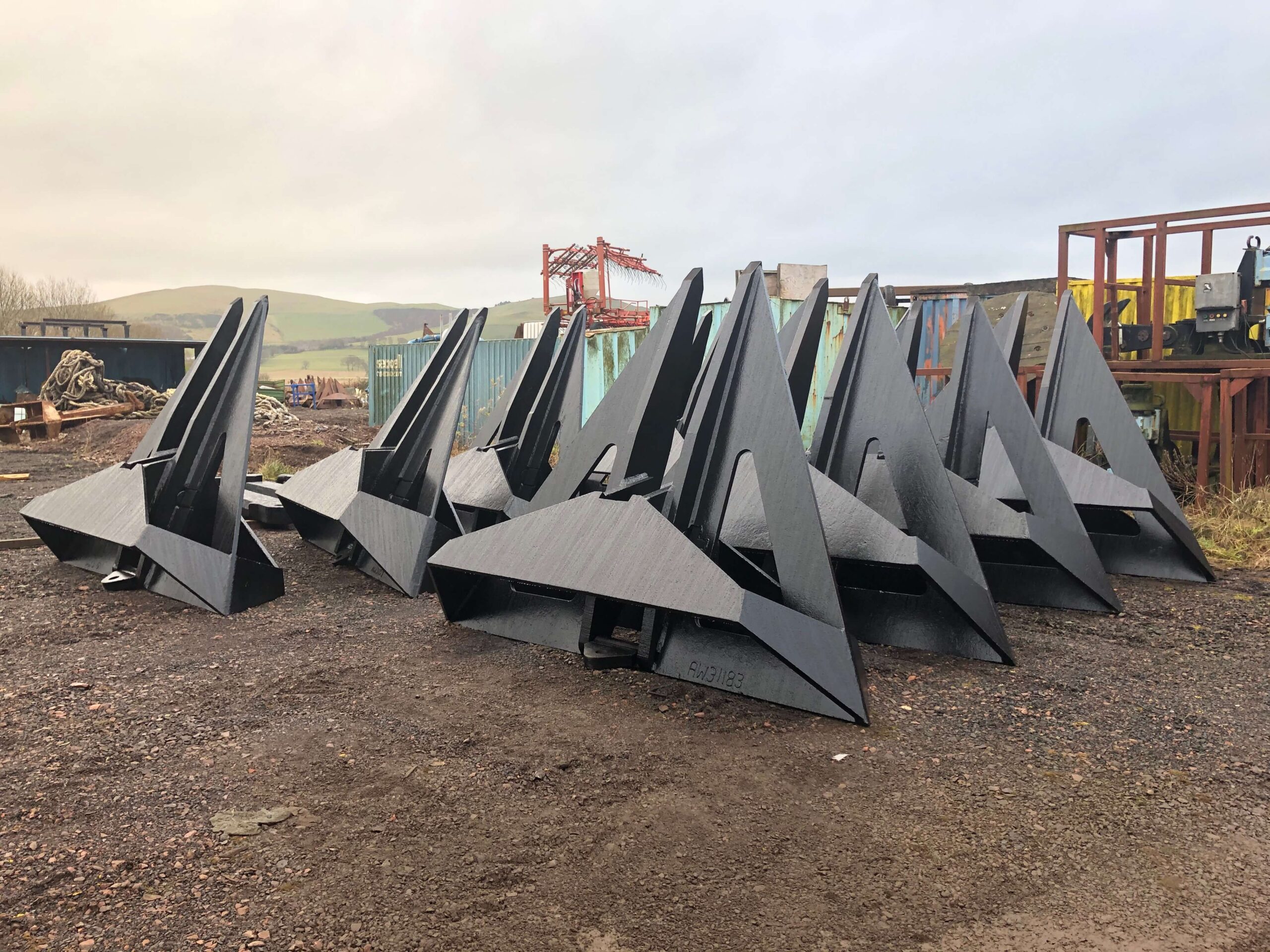 A pile of large ship anchors are stacked on a rocky ground in an industrial yard with shipping containers in the background.