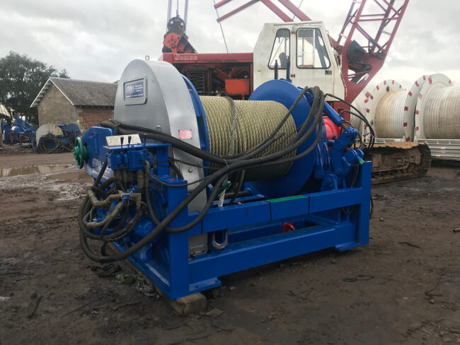 Blue industrial winch with thick yellow rope, surrounded by hydraulic cables, sits on muddy ground with a crane and barrels are in the background.
