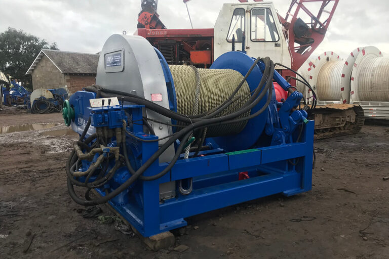 Blue industrial winch with thick yellow rope, surrounded by hydraulic cables, sits on muddy ground with a crane and barrels are in the background.